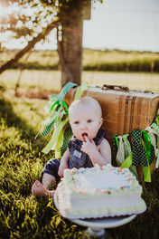 baby eats first birthday cake with his hands out in a summer field with vintage suitcase behind him