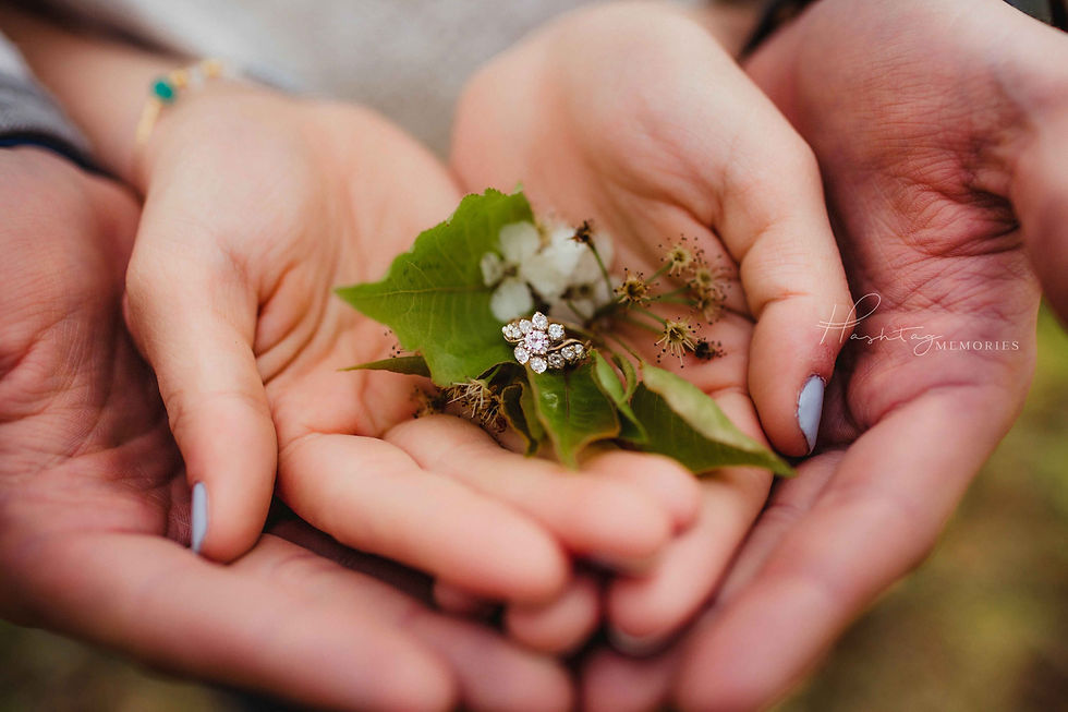 floral diamond engagement ring sits in couple's hands during engagement phot session. Ring sits on top of flower pear tree leave. Bride to be's finger nails painted periwinkly, hands are stacked with his on bottom and hers on top.