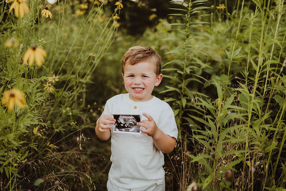 Smiling young boy in a white shirt holds an ultrasound photo amid tall wildflowers; faint watermark reads Happily Memories.