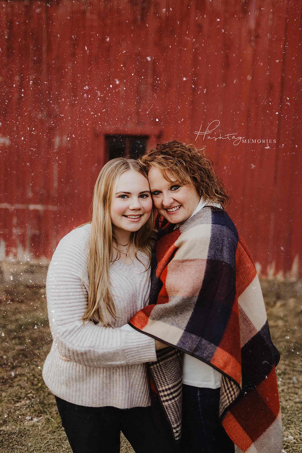 Two smiling women hug in front of a red barn, wrapped in a plaid blanket. Snow falls, creating a warm, cozy atmosphere. Text reads "Hashtag Memories."