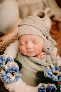 baby in grey sleepy hat holds teddy bear in wooden bucket, wearing green swaddle wrap with blue spring flowers in photography studio