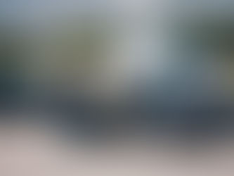 Mann öffnet Tür eines grauen Porsche-Cabrios für Fahrer. Schwarzer Porsche und weißer Hubschrauber im Hintergrund. Sonniger Tag.