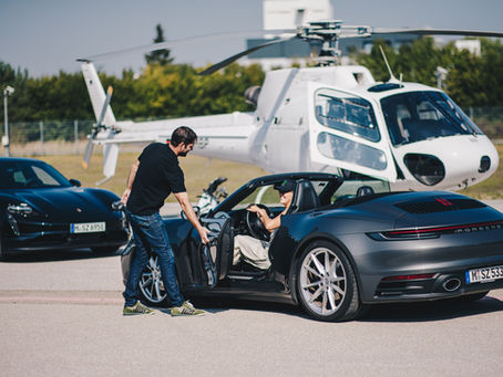 Mann öffnet Tür eines grauen Porsche-Cabrios für Fahrer. Schwarzer Porsche und weißer Hubschrauber im Hintergrund. Sonniger Tag.