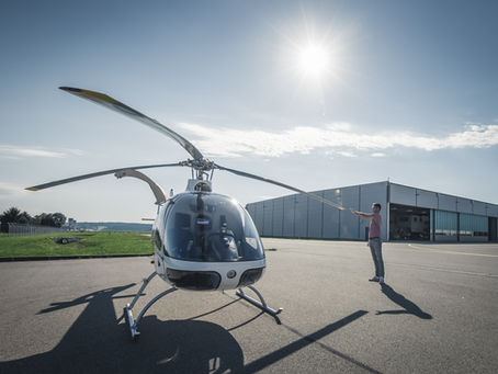 Ein Mann steht auf einem Flugfeld bei Sonnenschein neben einem weißen Hubschrauber, vor einem großen Hangar.