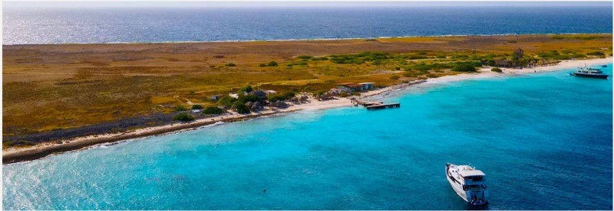Panoramic aerial shot of Klein Curaçao’s flat, uninhabited terrain—golden scrublands behind a narrow strip of white beach—with two excursion boats anchored in the clear, shallow blue sea.