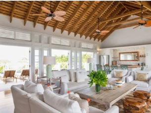 A bright, airy living room with soaring exposed-wood vaulted ceilings and three tropical-style ceiling fans. Two white slipcovered sofas and wooden stump stools surround a rustic coffee table, while floor-to-ceiling French doors and transom windows flood the space with light and open onto a shaded terrace. In the background, a breakfast bar with green wicker stools and a glimpse of a bedroom complete the open-concept, island-inspired interior.