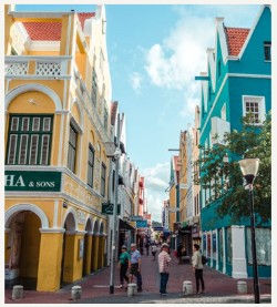 A narrow pedestrian street in Willemstad’s historic Punda district, flanked by vibrant, Dutch colonial–style buildings painted yellow, teal and white. Ornate gabled rooftops rise above arched doorways and shuttered windows, while a small group of people strolls and chats under a bright blue sky.