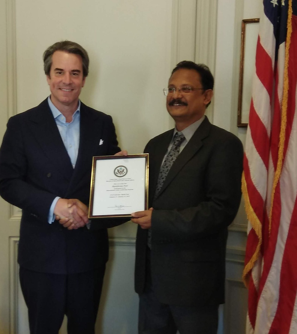 Two men in suits shake hands, one holding a framed certificate. They're smiling, with an American flag in the background. Formal setting.