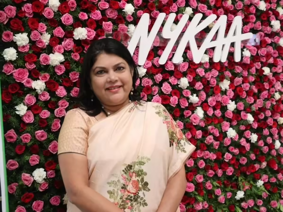 Smiling woman in a floral saree stands before a vibrant rose backdrop with "NYKAA" text. Background full of pink, red, and white roses.