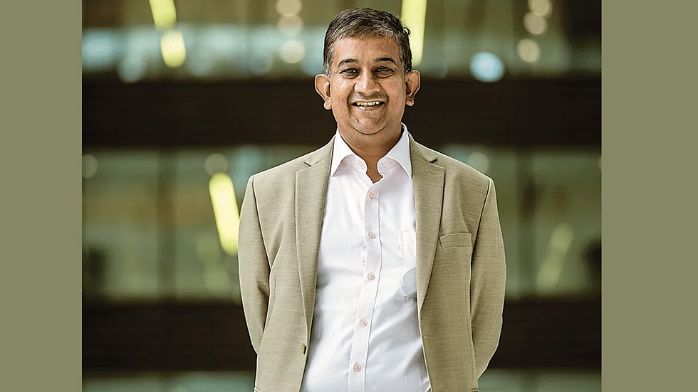 Smiling man in a beige suit and white shirt stands in front of a blurred modern building with glowing lights, conveying a confident mood.