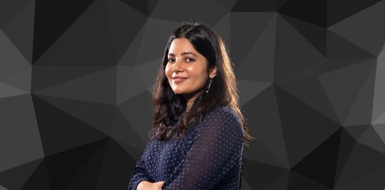 Woman with long dark hair and a navy blue polka dot top stands confidently against a geometric dark background.