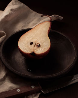 Pera cortada sobre plato oscuro con tela clara en una composición editorial de alimentos y objetos.