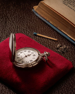 Reloj de bolsillo sobre tela roja con libro y cerillo en una escena editorial de objetos y memoria.