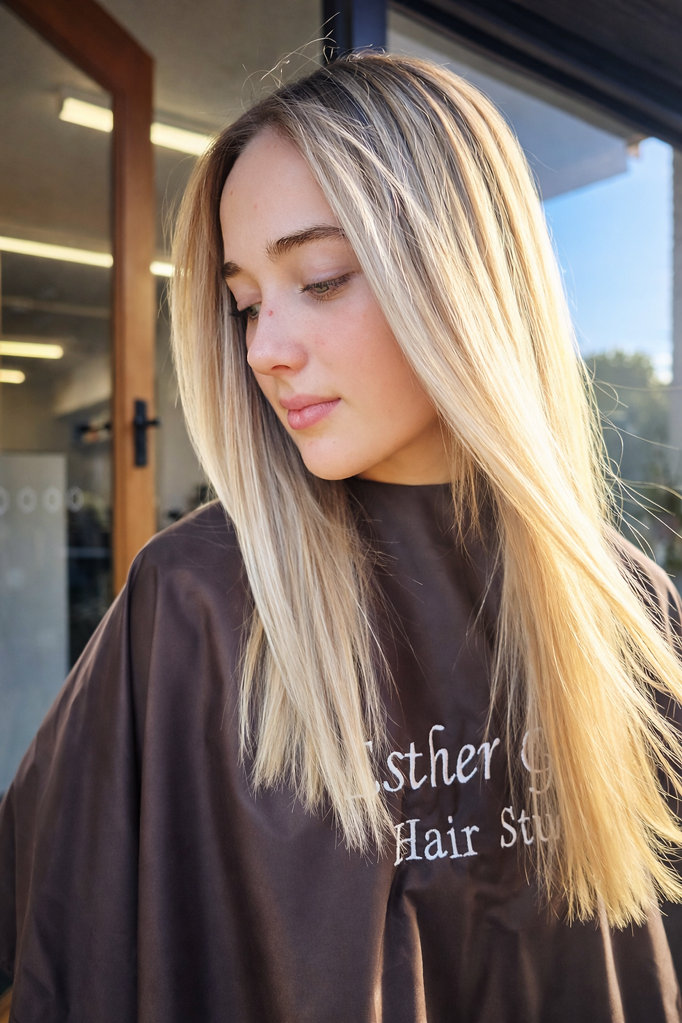 Young woman with long blonde hair in a salon, wearing a brown cape with visible esther grace hair studio text. Bright, natural light illuminates the scene.