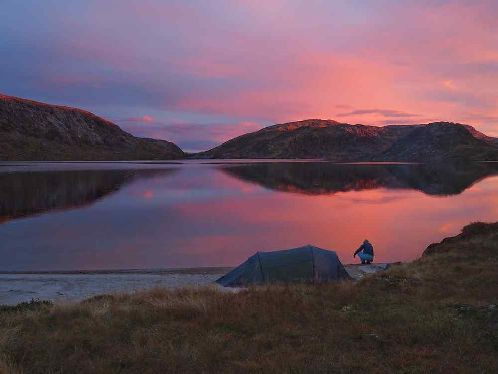Tent and crouching camper on a quiet lakeshore at pink sunset, with mountains reflected in calm water.