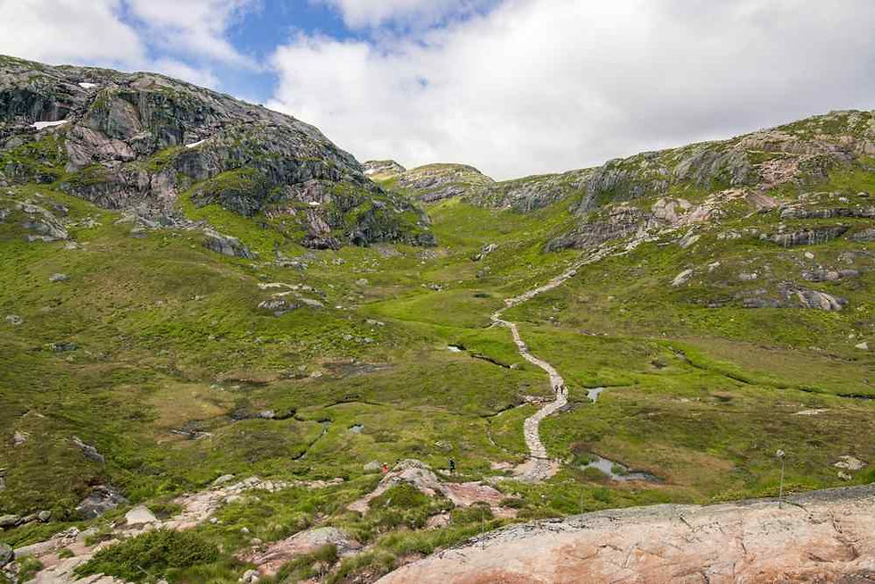 Hikers follow a winding stone path through green rocky mountains under a cloudy sky.