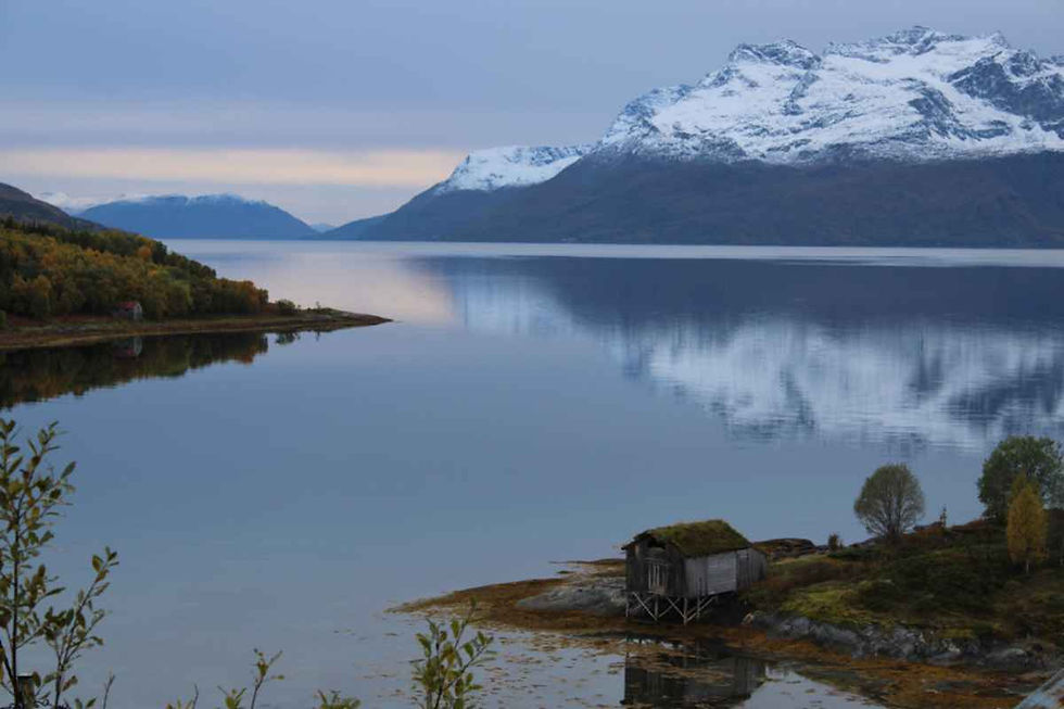 Elvelund by Gratangen fjord with a turf-roof hut and snowy peaks reflected in calm water.