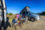 A large family enjoys a snack outside their camper in a designated camping area, setting up a small outdoor picnic with folding tables and chairs.