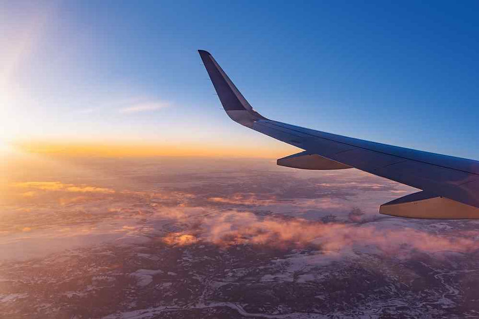 Airplane wing above clouds at sunset, with orange light and blue sky over a hazy landscape.