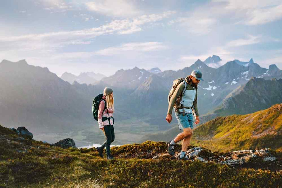 Two hikers with backpacks trek along a grassy mountain ridge at sunrise, with jagged peaks and cloudy sky behind.