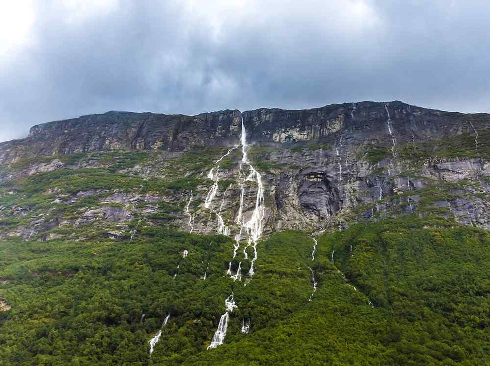 Multiple waterfalls cascade down a steep rocky cliff into lush green forest under a cloudy sky, creating a dramatic mountain scene