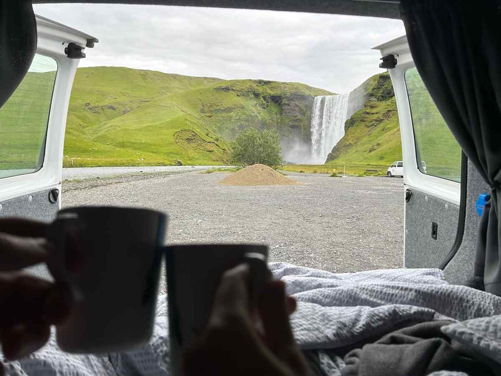 Dos tazas de café desde la cama de una camper, con las puertas traseras abiertas y una cascada al fondo en Islandia.
