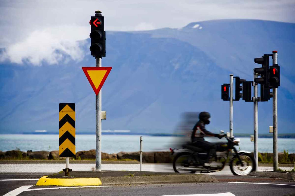 Un motorista y su moto en movimiento son captados a alta velocidad circulando por un tramo de una carretera islandesa con varias señales y semáforos, y una espectacular montaña detrás