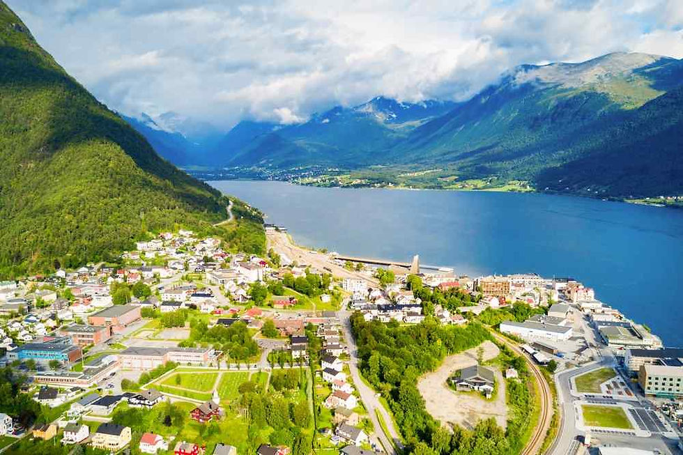 Aerial view of a lakeside town between green mountains and blue water under cloudy sky, with roads and buildings below.