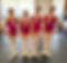 Four smiling ballet students wearing matching pink leotards, white tights, and ballet slippers, each with a numbered tag from 1 to 4, standing in a dance studio