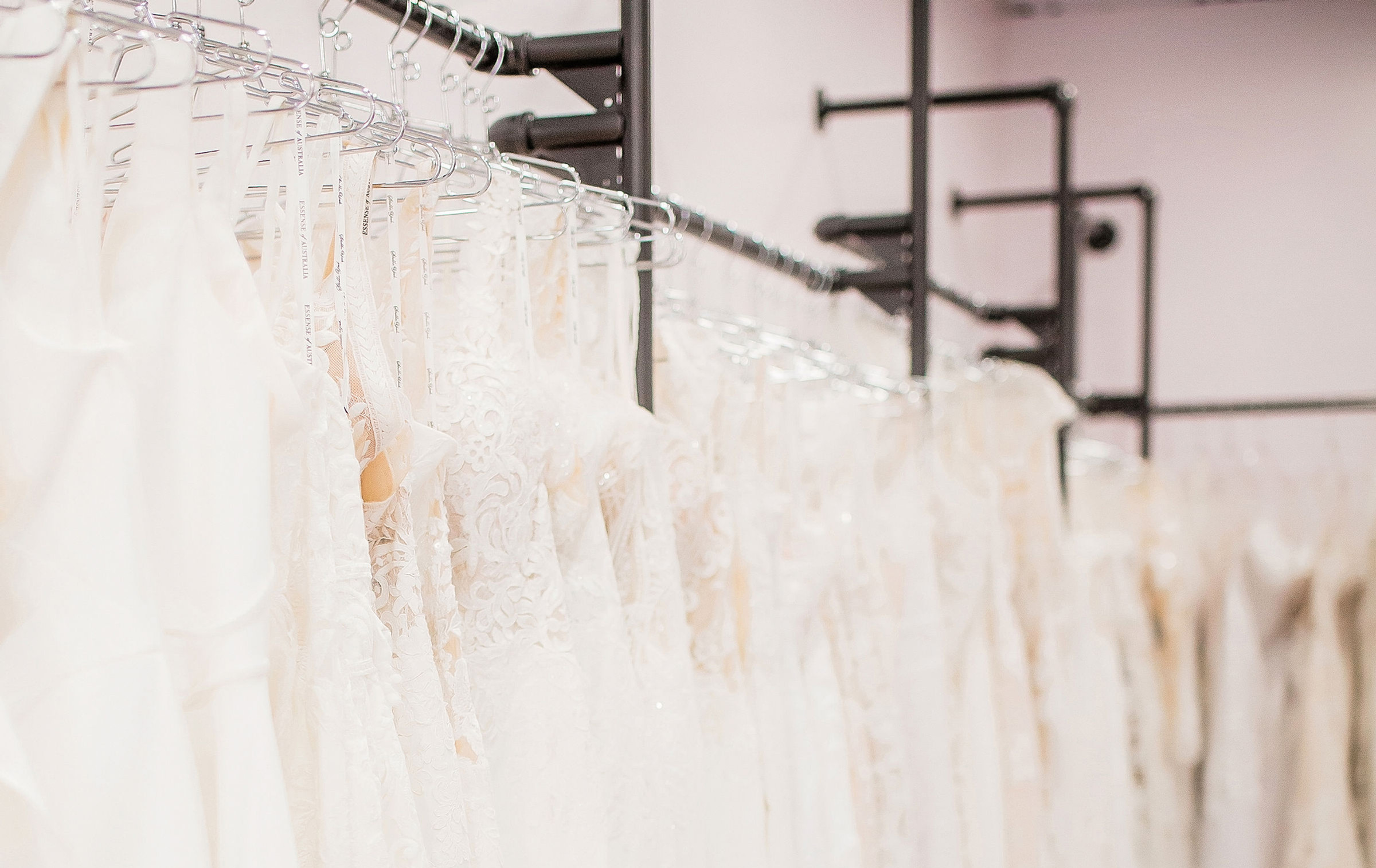 Rows of white wedding dresses on hangers in a bridal boutique, softly lit and elegant, with black rack frames in the background
