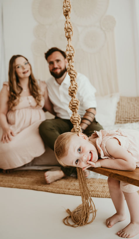 Ein fröhliches Kind auf einer Schaukel beim Babybauchshooting Hildesheim mit den Eltern im Hintergrund.