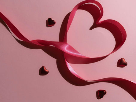 A pink background with a pink ribbon in the shape of a heart with heart-shaped chocolates in various places next to the ribbon-shaped heart