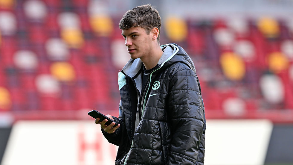 A person in a black jacket with a logo, holding a phone, stands in a blurred stadium setting with red and yellow seats.