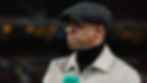 Man in glasses and gray flat cap, wearing a beige coat and black turtleneck, holds a teal microphone. Blurred background with warm lights.