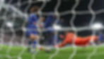 Chelsea players in blue and black jerseys compete near a goal, with a goalie in orange diving to save. Bright stadium lights and crowd in background.