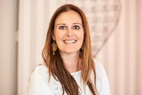 Lächelnde Frau mit langen braunen Haaren und weißem Oberteil vor hellem Hintergrund.