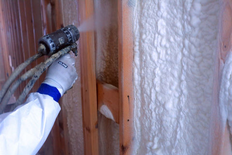 Hand with 'GLOVES' glove spraying foam insulation onto wooden wall studs.