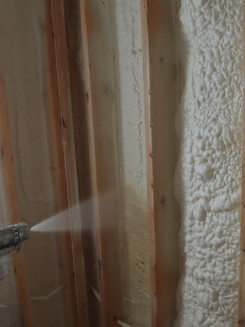 Person spraying foam insulation onto wooden wall studs for home construction.