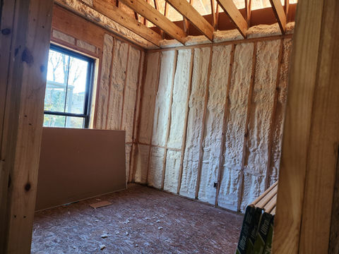 Unfinished room showing spray foam insulation on walls, wood framing, window.