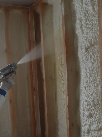Worker applying spray foam insulation to wooden wall studs near a window.