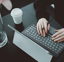 Ein Mensch, wo man nur die Hand sieht, der auf einem Laptop tippt. Neber dem Laptop steht ein Kaffeebecher und eine Pflanze mit Wasser am Boden.