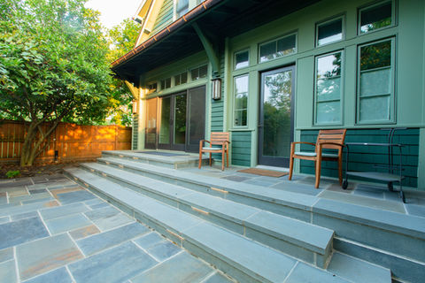 Green house exterior, stone patio, steps, chairs, and trees. Home.