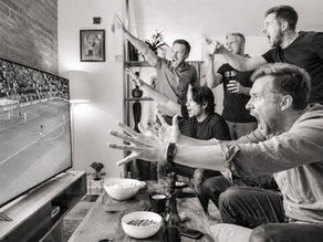 Zwart-witbeeld van een groep mannen die tijdens een voetbalwedstrijd boos en emotioneel reageert op wat er op het televisiescherm gebeurt.