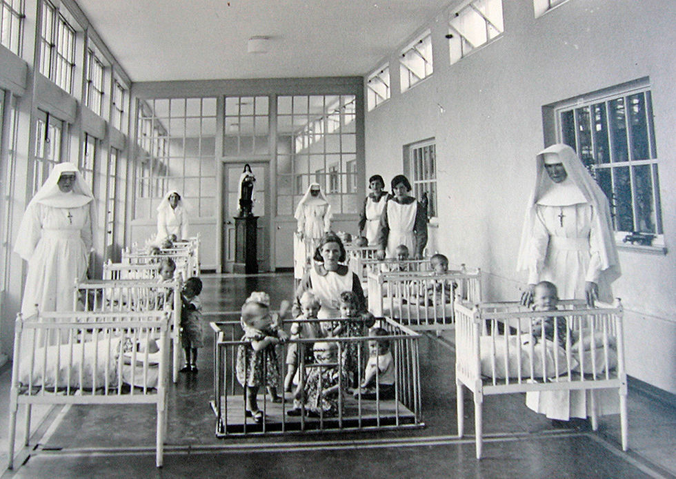 Historische opname van een kindertehuis waar nonnen en verpleegsters baby’s verzorgen in een steriele, afstandelijke omgeving — een beeld van zorg zonder aanraking.