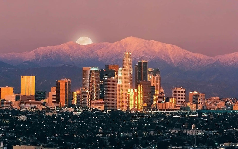 City skyline with illuminated buildings at sunset, snow-capped mountains, and a rising full moon in the background under a pink sky.