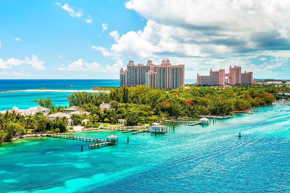 Tropical island with lush greenery, overwater bungalows, turquoise ocean, and distant pink resort buildings under a bright blue sky.