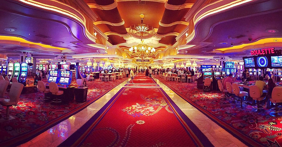 Casino interior with vibrant slot machines, red patterned carpet, and elegant chandeliers. People are gaming, with "ROULETTE" sign visible.