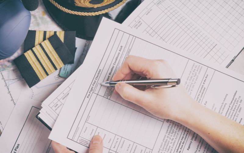 Hand filling out aviation documents with a pen. Pilot uniform details nearby. Papers spread on a table. Navy and gold accents prominent.