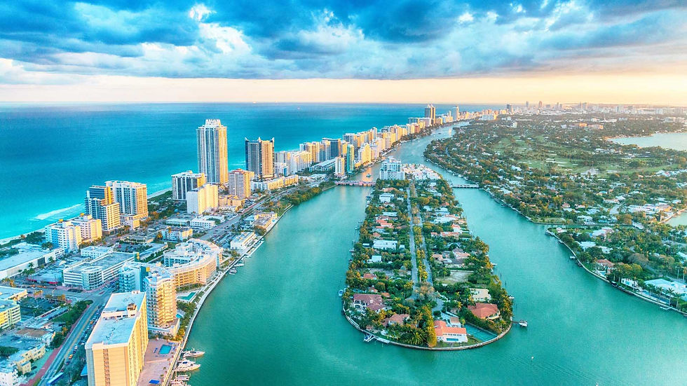 Aerial view of a coastal city with high-rise buildings, a winding canal, and lush greenery. The sea is vivid blue under a cloudy sky.