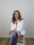 Smiling woman with light brown hair sitting on stool, wearing a white blazer and jeans
